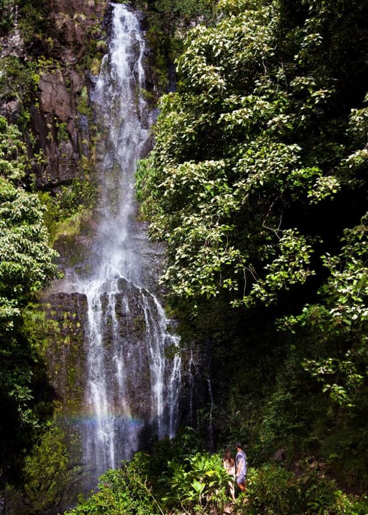 Two people stand near the bottom of Wailua Falls waterfall in Maui Hawaii.