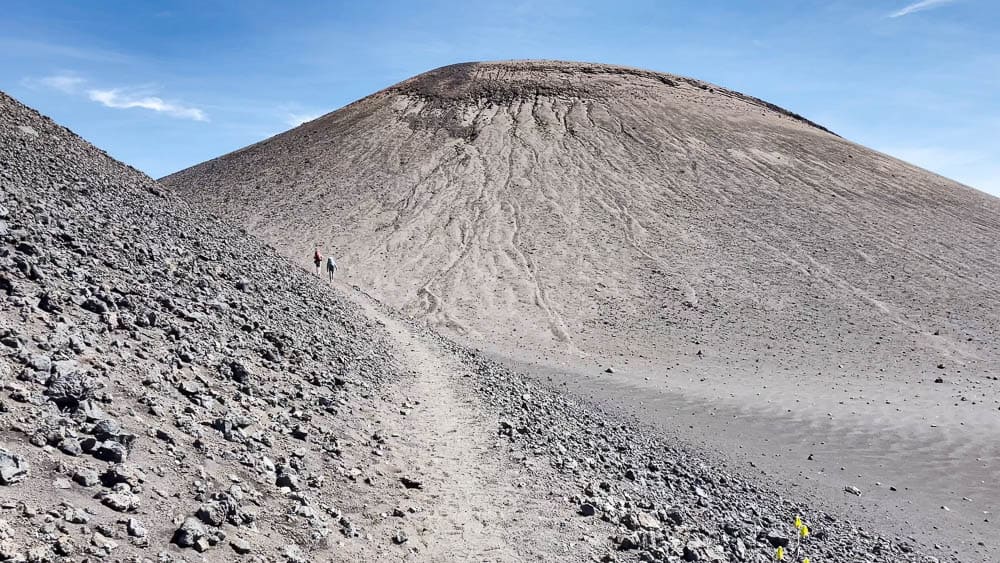 Two people walking in the distance on a barren gray hiking trail around a volcanic cinder cone.