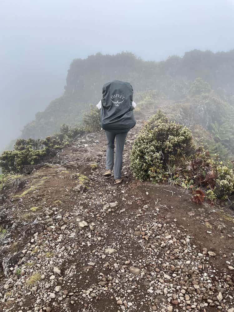 A hiker with a large pack and surrounded by fog on the rocky Halemauʻu trail portion called Rainbow Bridge. 