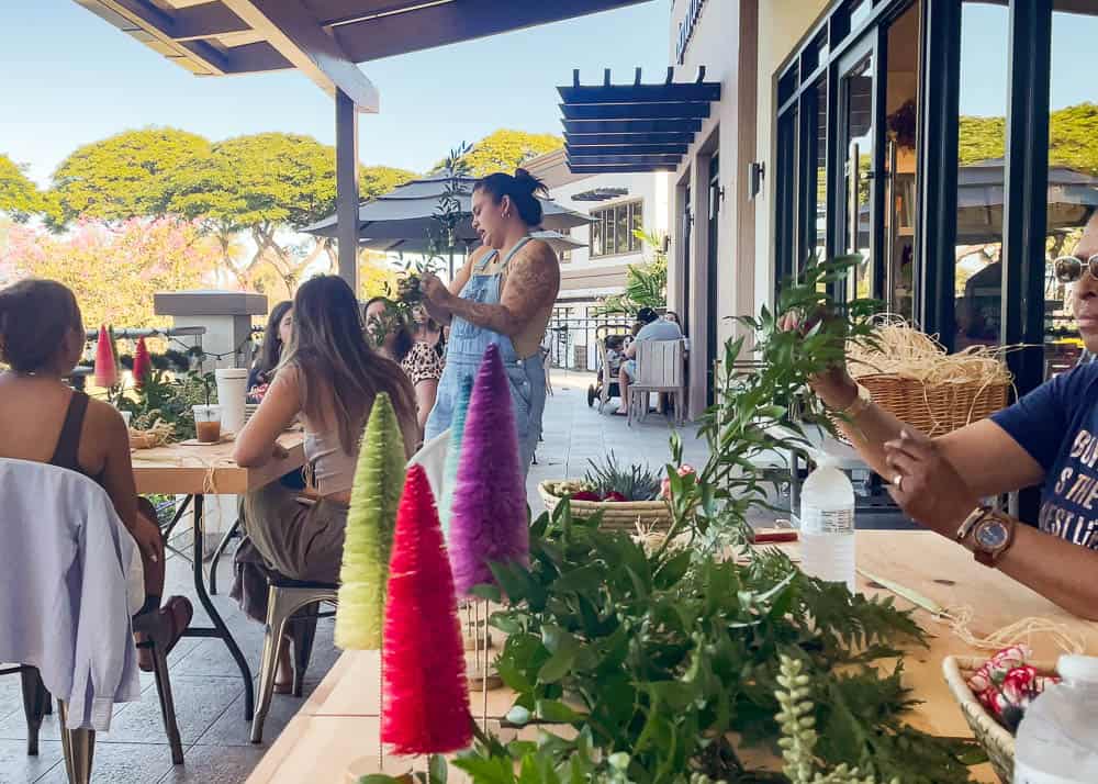 A lei making instructor holds up foliage while a woman picks up leaf supplies from the table at Paper Garden Maui