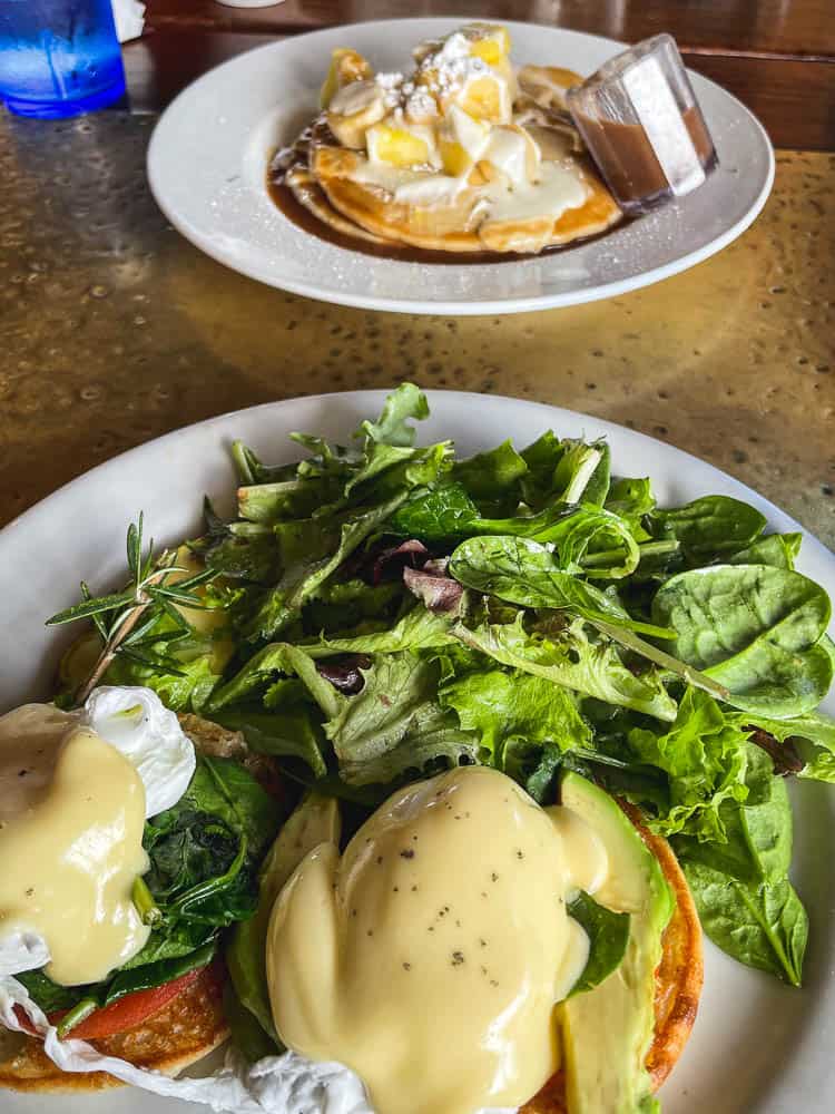 Eggs benedict and salad greens on a plate in the foreground. Pancakes topped with syrup and bananas on a plate in the background.