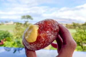 A hand holding a filled malasada from Sugar Beach Bake Shop with a pool, golf course, and sky in the background