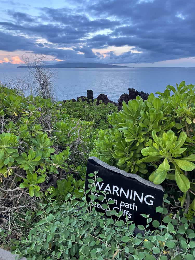 A stone warning sign telling pedestrians to stay on the path due to steep cliffs with the ocean and Molokini Crater in the distance