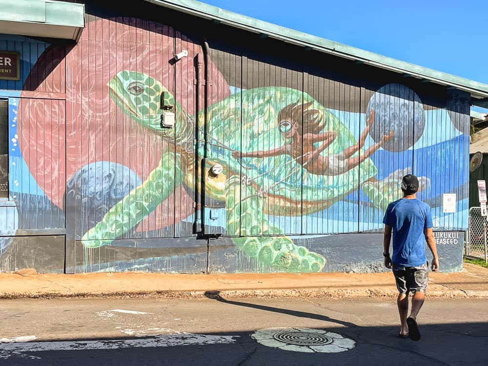 A man walking across the street near a mural of a sea turtle and swimmer on the side of a building in Wailuku, Maui